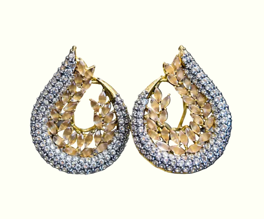 A pair of teardrop-shaped gold earrings paved with clear stones and featuring a central gold leaf pattern, displayed against a solid black background.