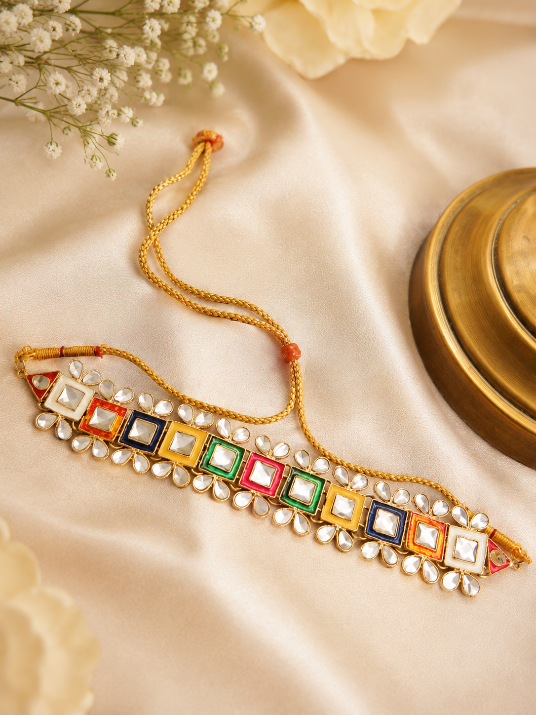 A gold-plated choker necklace with colorful square inlays (red, green, blue, orange) and white stones, decorated with small white pearls, displayed on a beige satin fabric background.